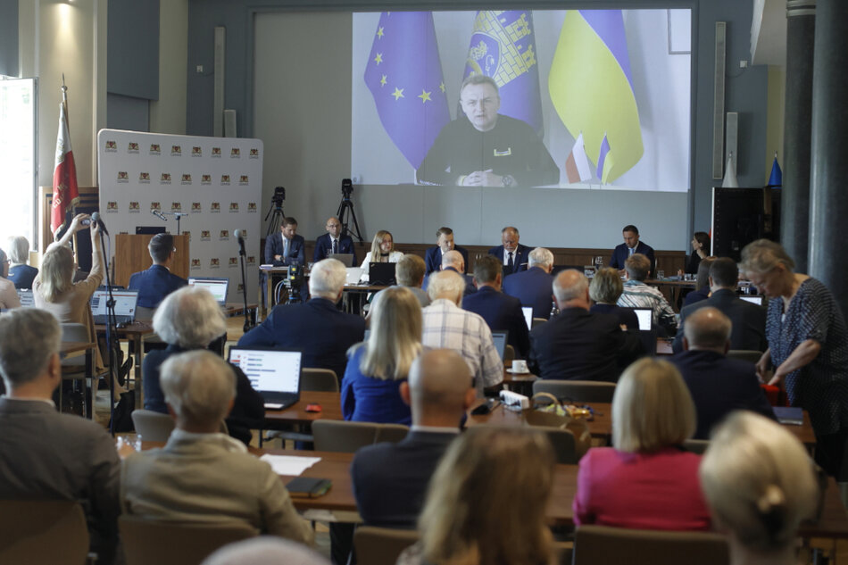 na pierwszym planie plecy radnych siedzących przy biurkach, w tle na ścianie wyświetlany obraz z projektora, na nim mer