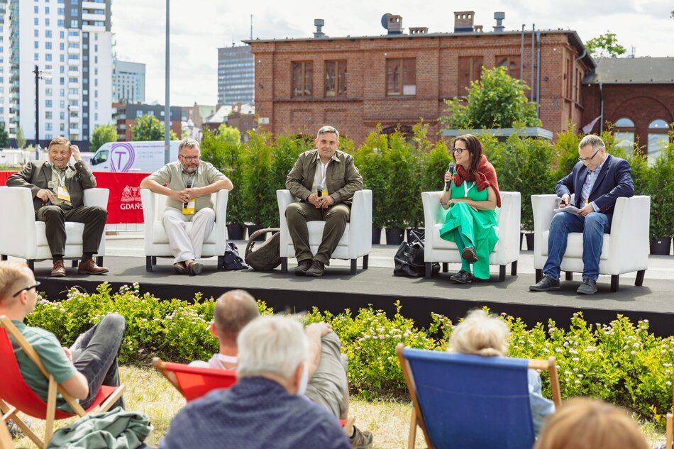na zdjęciu niewysoka scena na niej pięć jasnych foteli, na których siedzą i debatują osoby w średnim wieku, jedna kobieta i czterech mężczyzn