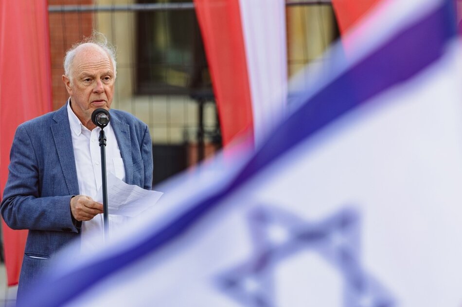 Starszy mężczyzna o siwych włosach stoi przemawiając przez mikrofon. Przed nim widać powiewającą niebiesko-białą flagę Izraela z Gwiazdą Dawida. Na prawo od niego jest flaga Polski