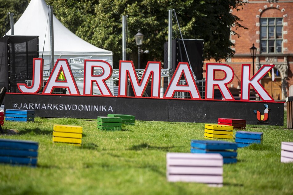 Duży napis JARMARK ustawiony na trawniku. Przed nim rozstawione kolorowe skrzynki