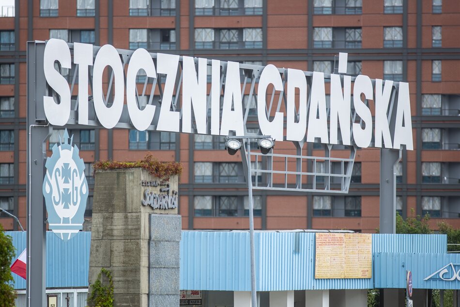 na zdjęciu duży biały napis stocznia gdańska, w tle fragment brązowego ceglanego budynku z dużą ilością okien