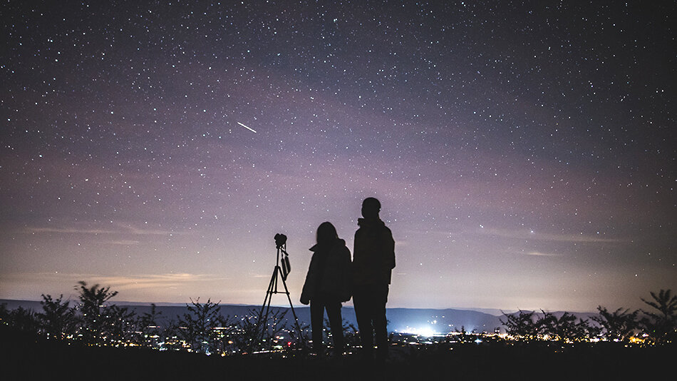 sylwetki dwojga ludzi z aparatem na statywie na tle gwieździstego nieba 