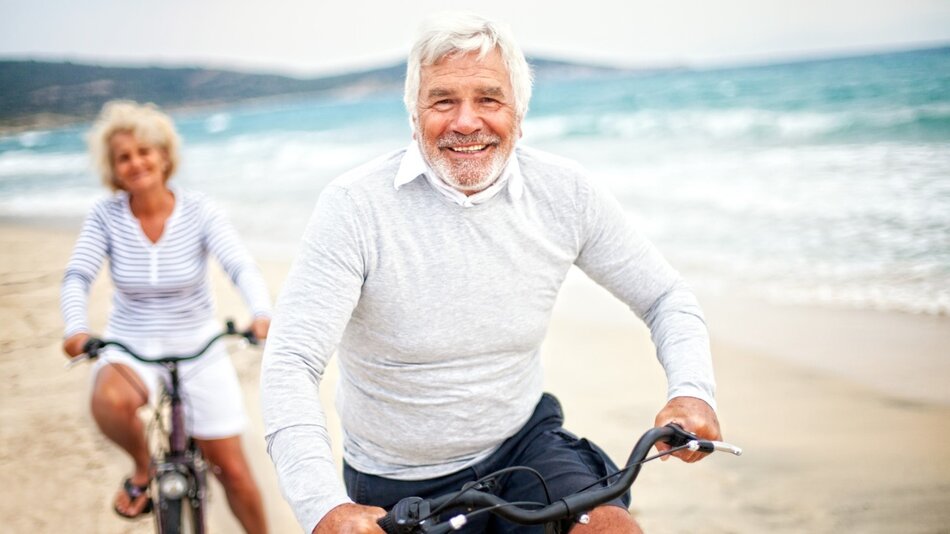 Plaża, siwy mężczyzna na rowerze, za nim kobieta na rowerze 