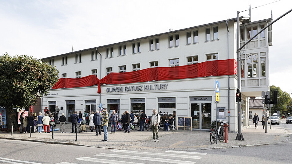 dwupiętrowy, biały budynek, otoczony zielenią, przed nim chodnik i ulica