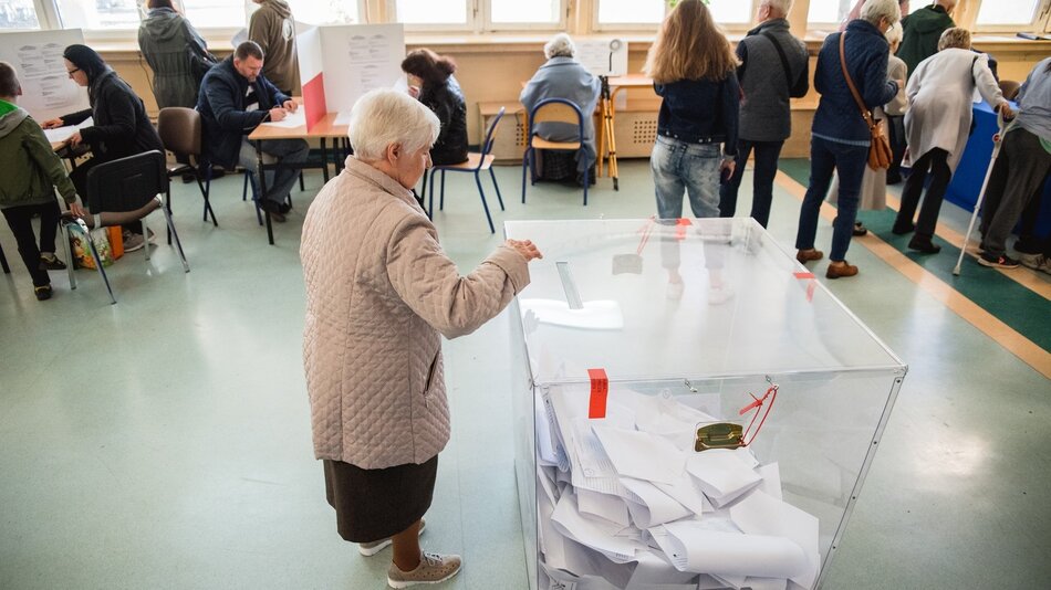 Lokal wyborczy, w którym starsza kobieta na tle innych osób wrzuca do urny kartę do głosowania