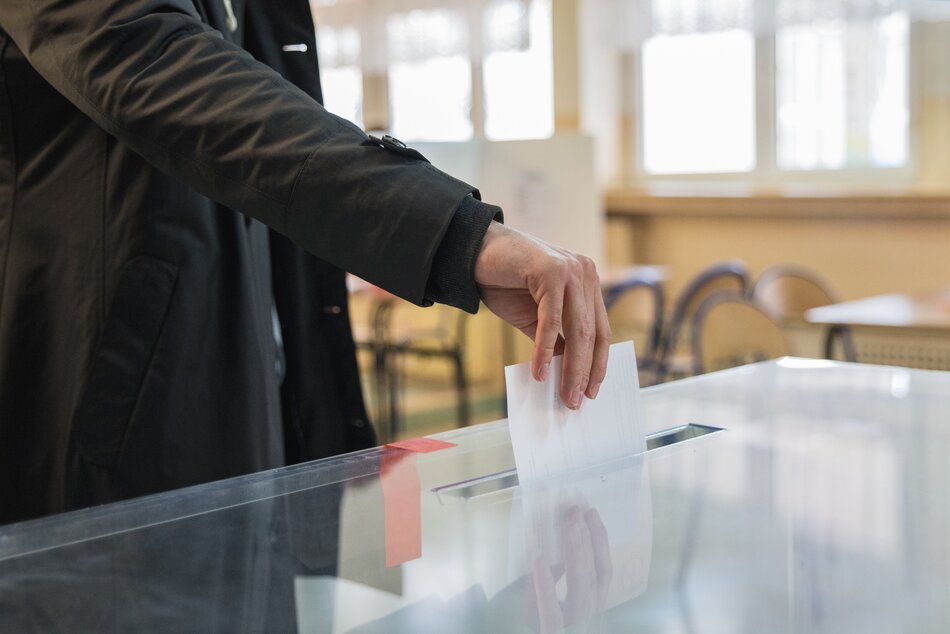 Zdjęcie przedstawia dłoń, która wrzuca kartkę wyborczą do podłużnego otworu w górnej ścianie urny wyborczej, która jest wykonana z przeźroczystego materiału 
