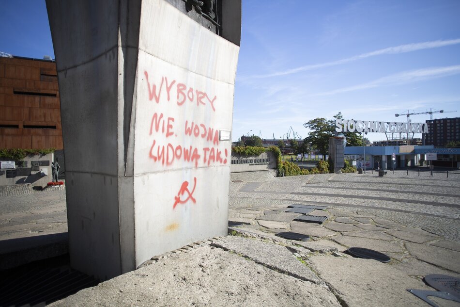 fragment stalowego pomnika z widocznym napisem namalowanym czerwoną farbą, w tle płot i brama wjazdowa 