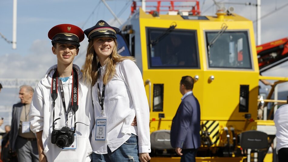 Dwie uśmiechnięte młode osoby w czapkach kolejarskich na tle pociągu