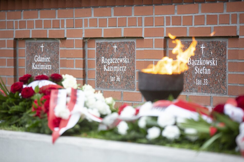 Zdjęcie przedstawia trzy imienne płyty nagrobkowe w ścianie Cmentarza na Zaspie. Pod płytami widzimy ułożone kwiaty z biało-czerwonymi wstęgami oraz znicz płonący żywym płomieniem 