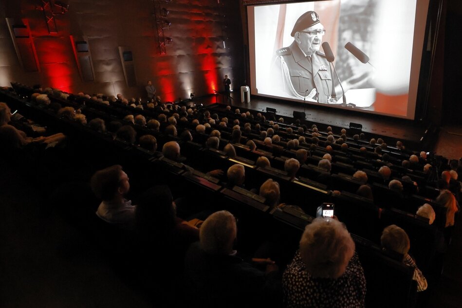 Sala koncertowa - widownia wypełniona, na scenie ekran, na którym wyświetla się zdjęcie starszego mężczyzny w mundurze.