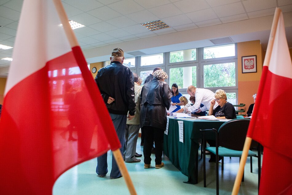 kolejka w sali, ludzie głosują za stołem komisja wyborcza, obok flagi biało-czerwnoe