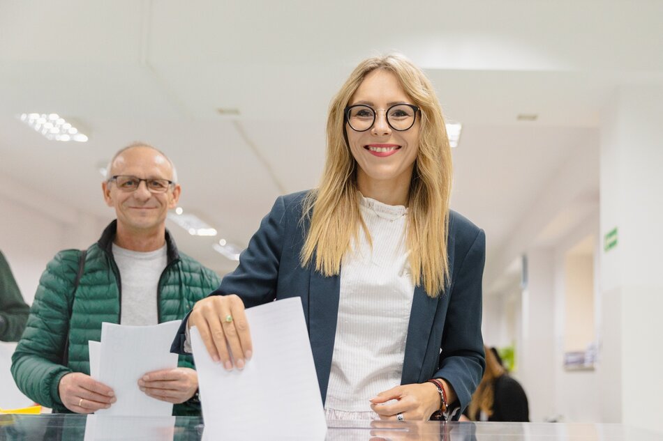 Uśmiechnięta młoda blondynka o długich włosach wrzuca karty wyborcze do urny do głosowania