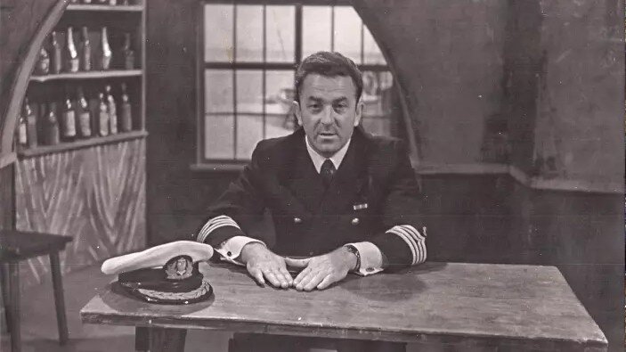 Czarno-białe zdjęcie: mężczyzna w marynarskim mundurze siedzi za stołem. Na stole leży czapka marynarska