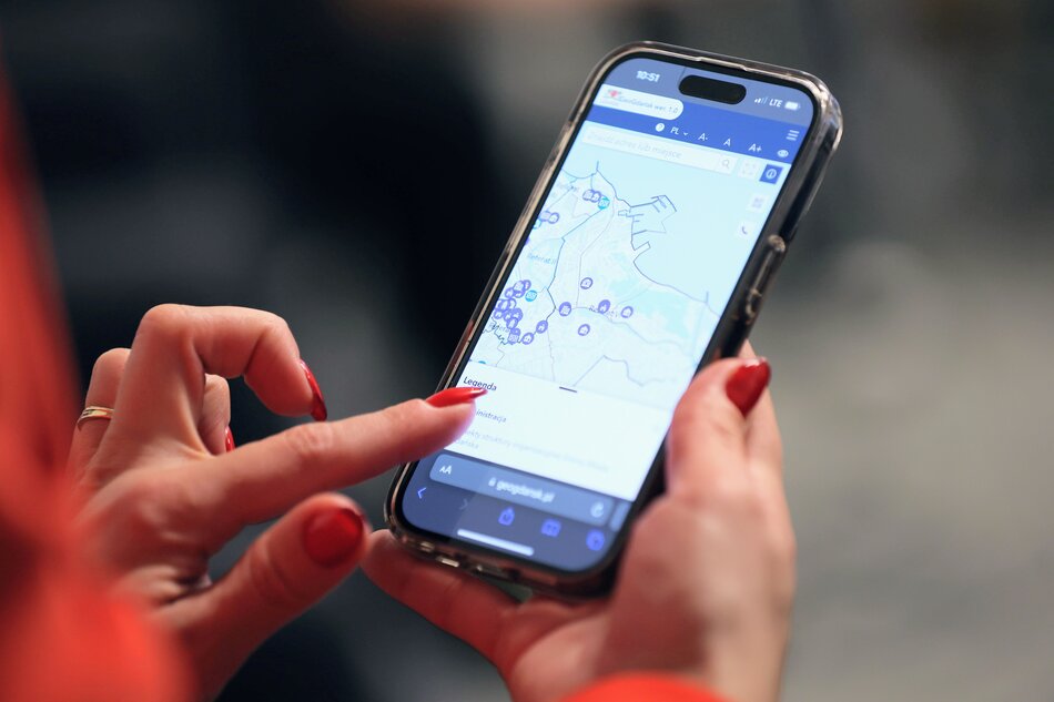 na zdjęciu smartfon, trzymany w kobiecej dłoni z wymalowanymi czerwonymi paznokciami
