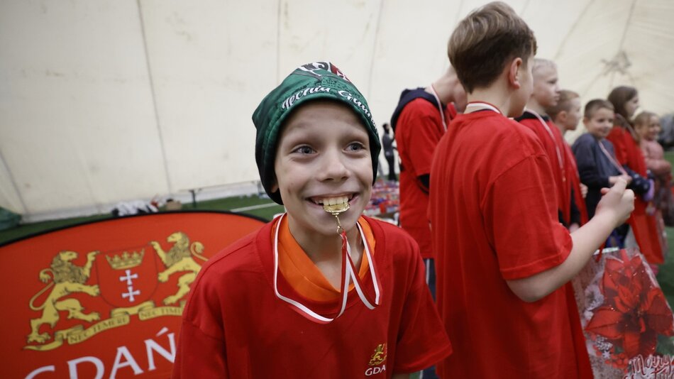 Uśmiechnięty chłopiec w czapce gryzący w ustach medal, obok niego inne dzieci