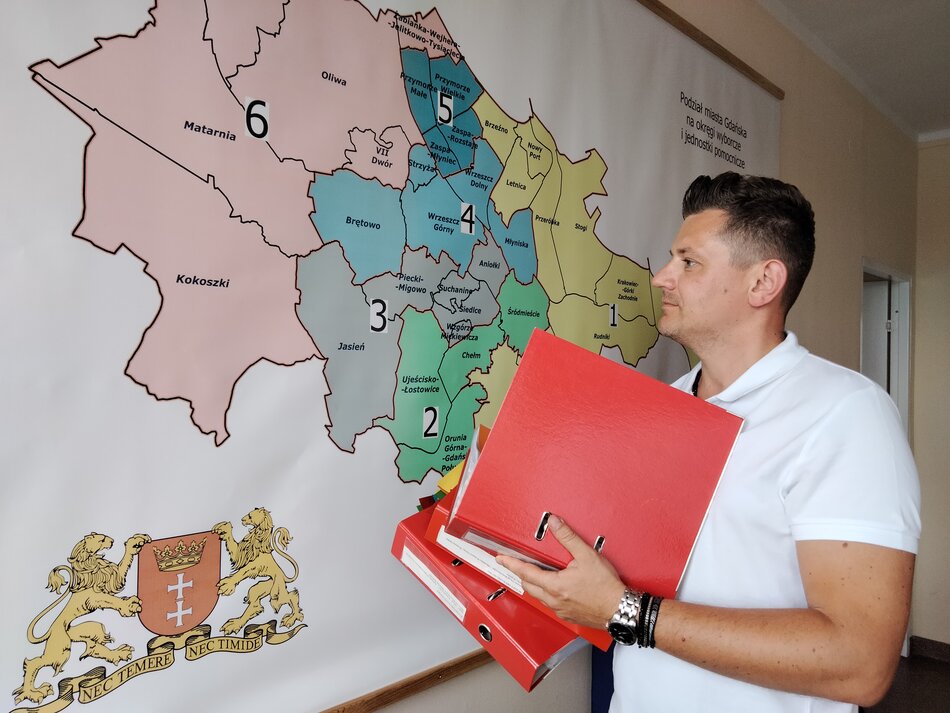 mężczyzna stojący z czerwoną teczką na tle mapy dzielnic miasta Gdańska