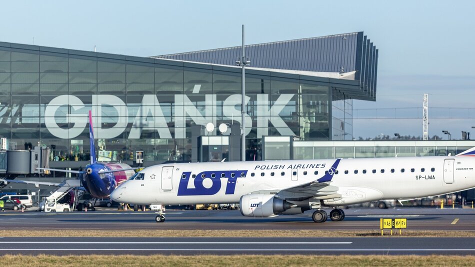 Na zdjęciu widać samolot pasażerski, który stoi na pasie startowym lotniska. Samolot jest pomalowany na biało z czerwonymi i niebieskimi paskami. Na kadłubie samolotu widnieje napis „Gdańsk” oraz logo LOT Polish Airlines.