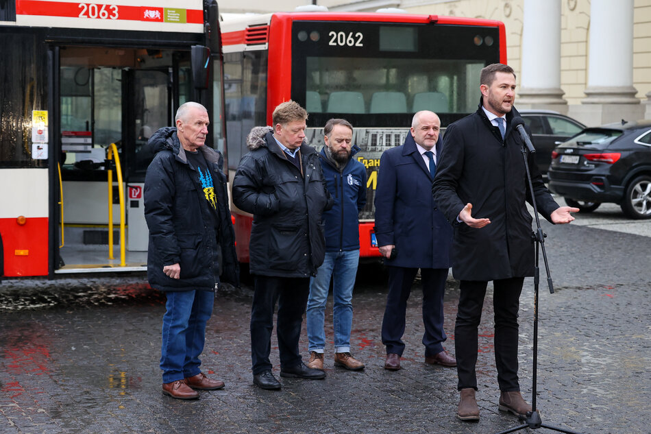 Pięciu mężczyzn w kurtkach stoi przed autobusem na placu. Najwyższy (Piotr Grzelak) przed mikrofonem