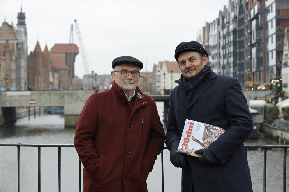 Na zdjęciu widać dwóch mężczyzn stojących na moście na rzece Motławie. W tle miejska architektura. Mężczyzna po lewej stronie jest ubrany w bordowy płaszcz i ma na głowie czarną, płaską czapkę. Noszi okulary i ma brodę z siwymi włosami. Drugi mężczyzna po prawej stronie ma na sobie ciemnoniebieski płaszcz i również czarną, płaską czapkę. Trzyma czasopismo 30 Dni z kolorową okładką. Za nimi widać rzekę, most, a także ciekawe, kolorowe budynki, które są częścią starego miasta. Na zdjęciu jest pochmurno i wygląda na zimno.