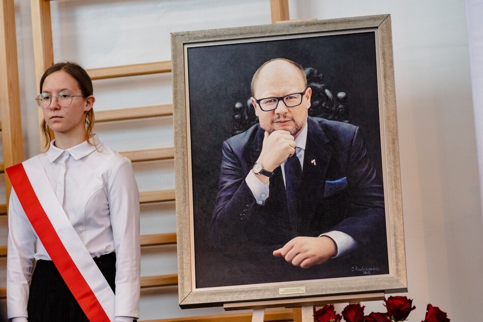 ubrana na galowo uczennica przepasana szarfą z flagą Polski pozuje obok portretu śp. prezydenta - uśmiechniętego mężczyzny w okularach
