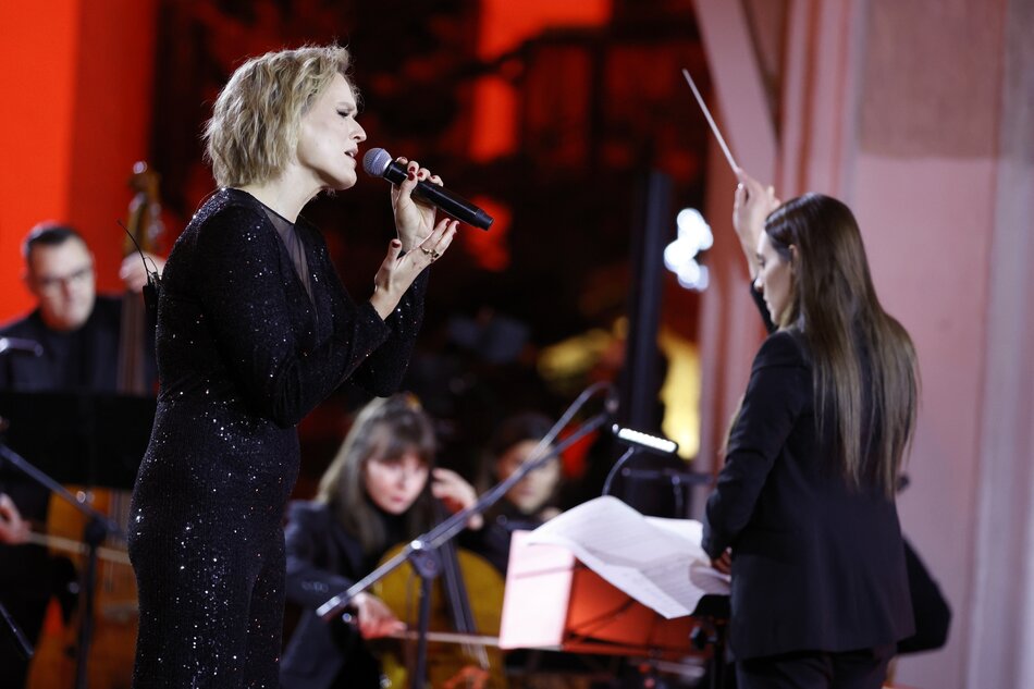 na zdjęciu artystka śpiewająca do mikrofonu, ma czarną sukienkę, w tle dyrygentka wymachująca rękoma i muzycy orkiestry