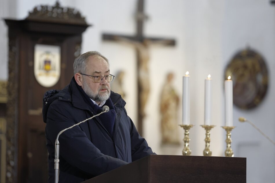na zdjęciu starszy mężczyzna solidnej postury z siwymi włosami i brodą, w okularach, stoi za pulpitem, mówi do mikrofonu, po prawej widać kościelne msze na ołtarzu i fragment ołtarza
