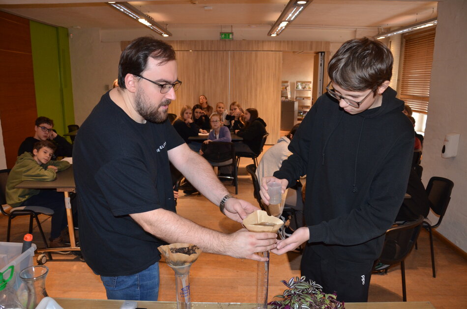 mężczyzna i nastolatek przy stole, na którym stoją próbówki, na jednej z nich filtr, przelewają przez niego jakiś płyn, w tle przygląda się grupa nastolatków siedząca przy stołach