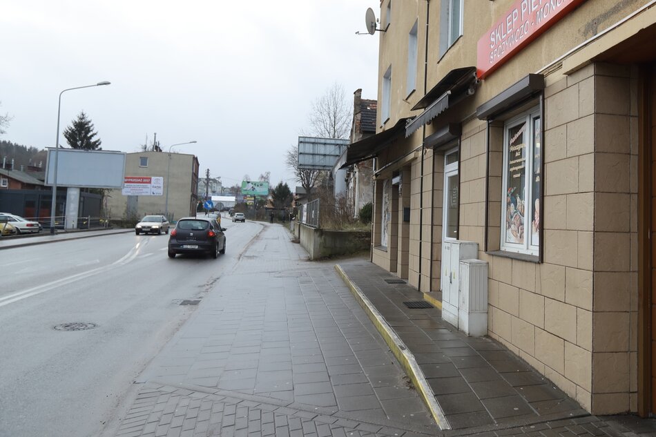 na zdjęciu po lewej fragment ulicy i chodnika, ulicą jedzie samochód osobowy, po prawej fragment budynku o kolorze piaskowym