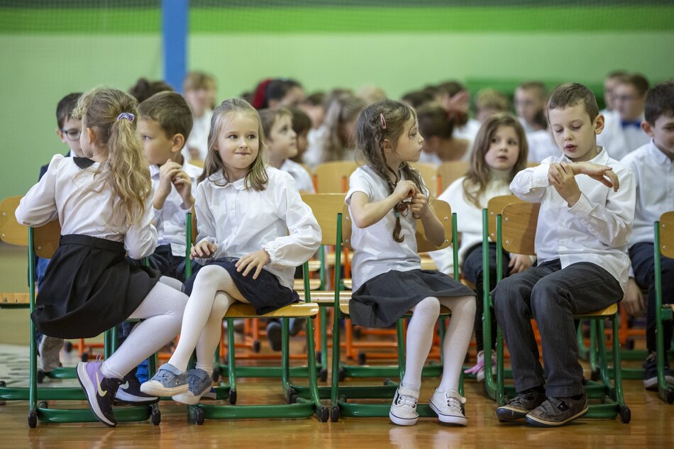 dzieci w wieku ok 6-8 lat, chłopcy i dziewczynki w galowych strojach, siedzą w ławkach na sali gimnastycznej