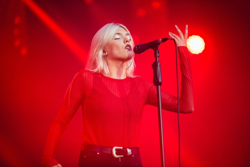 szczupła blondynka śpiewa do mikrofonu na scenie skąpanej w czerwonym świetle