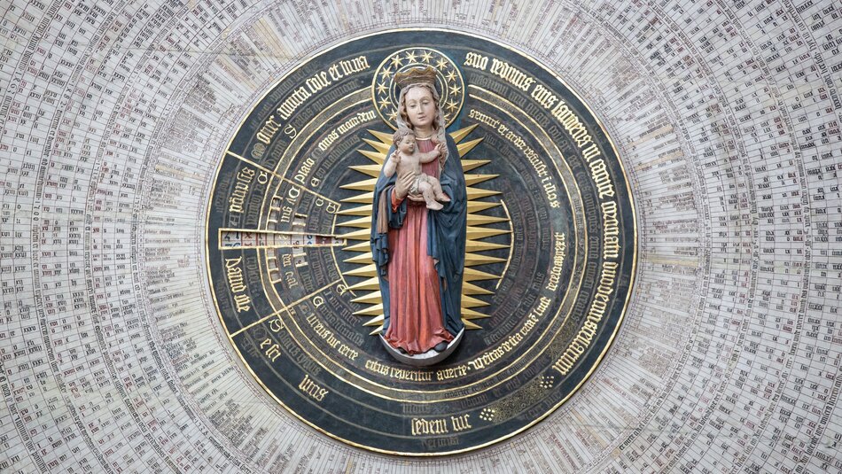 Zdjęcie przedstawia szczegół astronomicznego zegara w formie astrolabium, który jest bogato zdobiony. Na pierwszym planie widoczna jest trójwymiarowa rzeźba przedstawiająca postać kobiecą, najprawdopodobniej Matkę Boską z Dzieciątkiem Jezus na ręku. Rzeźba jest kolorowa – postać kobieca ma na sobie niebieską szatę z czerwonym elementem odzieży pod spodem, a Dzieciątko Jezus jest nagie. Wokół tej figury rozmieszczone są złote promienie, tworzące efekt aureoli. W tle znajduje się właściwa tarcza zegara, wykonana w kolorach złota i czerni, z licznymi napisami i znakami. Zegar składa się z koncentrycznych kręgów z różnymi cyframi rzymskimi i arabskimi, tekstami w języku łacińskim, a także wskazówkami na poszczególne dni, miesiące i znaki zodiaku. Wewnętrzne kręgi są podzielone na segmenty, które prawdopodobnie odnoszą się do ruchów ciał niebieskich i ich wpływu na czas oraz daty.