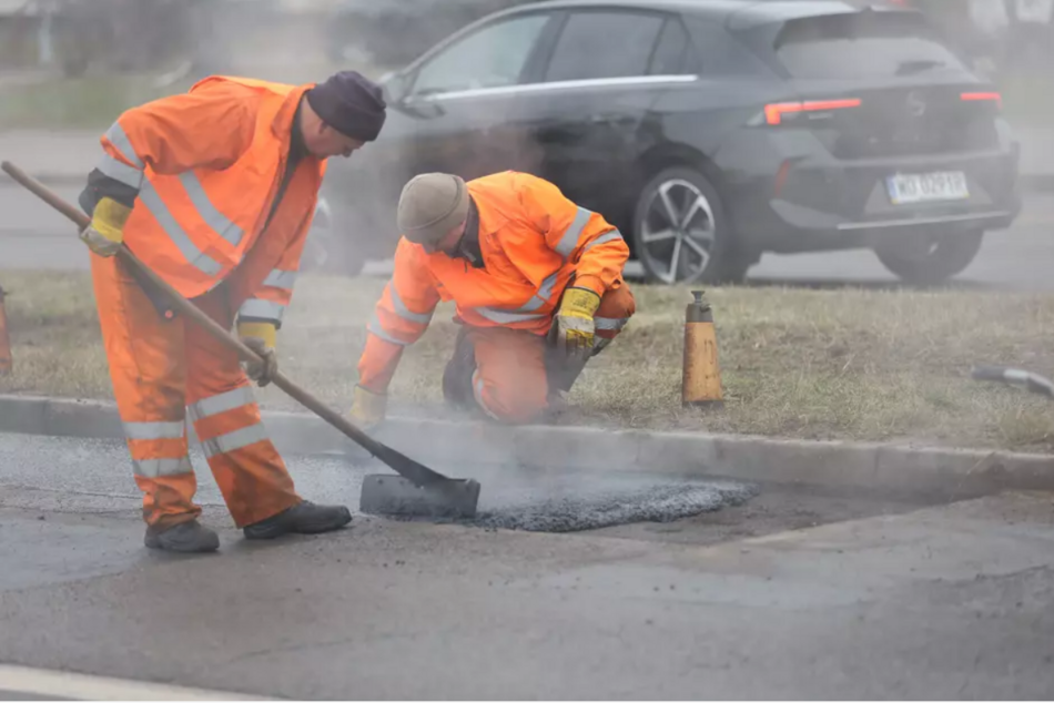 na zdjęciu dwóch pracowników w pomarańczowych robotniczych kombinezonach, jeden kuca przy jezdni, drugi rozprowadza łopatą gorący asfalt