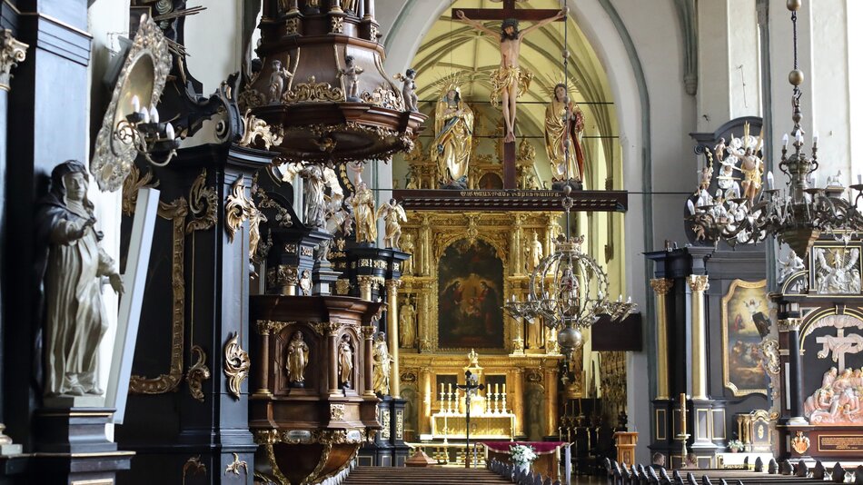 Zdjęcie przedstawia wnętrze kościoła o bogato zdobionej architekturze barokowej. Centralnym punktem jest złoty ołtarz główny z dużym, ciemnym obrazem w centrum, który jest otoczony przez liczne figury świętych i aniołów. Nad ołtarzem wisi duży krucyfiks, z Jezusem Chrystusem na krzyżu, a po jego bokach znajdują się figury świętych. Wzdłuż bocznych ścian ustawione są ciemne, drewniane konfesjonały z ozdobnymi elementami, w tym złotymi liśćmi i rzeźbionymi detalami. Widoczne są również liczne świeczniki, kandelabry i inne elementy liturgiczne. W tle można dostrzec sklepienie kościoła z łukami, co dodaje wnętrzu wrażenie przestronności i majestatu. Ławki dla wiernych są ustawione wzdłuż nawy, wskazując na funkcję sakralną tego miejsca. Ogólna kompozycja obrazu jest symetryczna, co podkreśla uroczysty i sakralny charakter kościoła