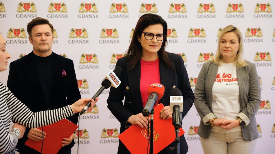Widzę zdjęcie, na którym trzy osoby uczestniczą w konferencji prasowej. Kobieta w centrum stoi za mównicą, ubrana jest w ciemny żakiet, czerwony sweter i ma na sobie okulary. Po jej prawej stronie stoi mężczyzna w czarnym golfie, a po lewej inna kobieta w szarym żakiecie i białej koszulce z czerwonym napisem. Wszyscy trzej wyglądają na zaangażowanych w to, co się dzieje, a kobieta w środku wydaje się być główną osobą przemawiającą. W tle widać logo, które wygląda na herb lub logo miasta, a mikrofony przedstawiają różne stacje telewizyjne i radiowe