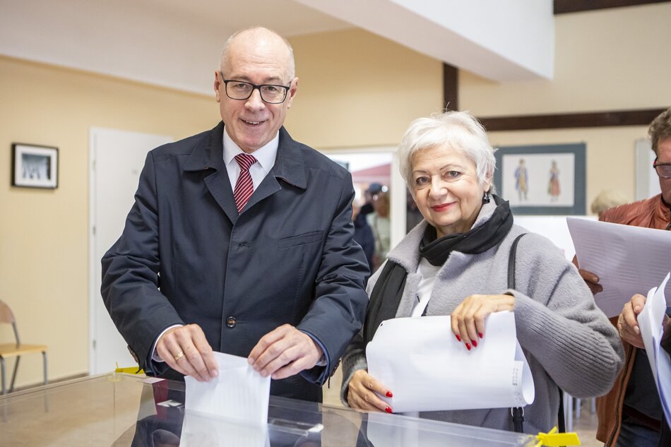 Mężczyzna i kobieta, oboje w wieku ponad lat 60, pozują do zdjęcia przy urnie wyborczej. Mężczyzna jest po lewej 
