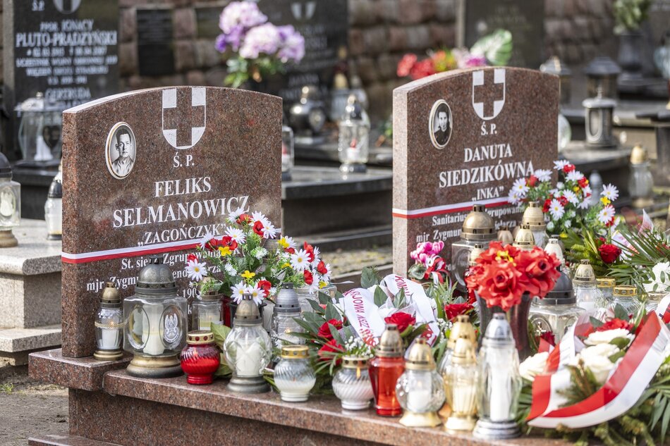 Dwa kamienne groby, znajdujące się obok siebie. Na nich liczne wieńce i kwiaty z biało-czerwonymi wstążkami 