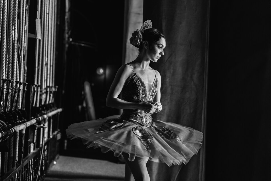 Tancerka stoi w kulisie, ma lekko pochyloną głowę, jest zamyślona. Jest ubrana w strój baletowy, sukienkę i tutu.