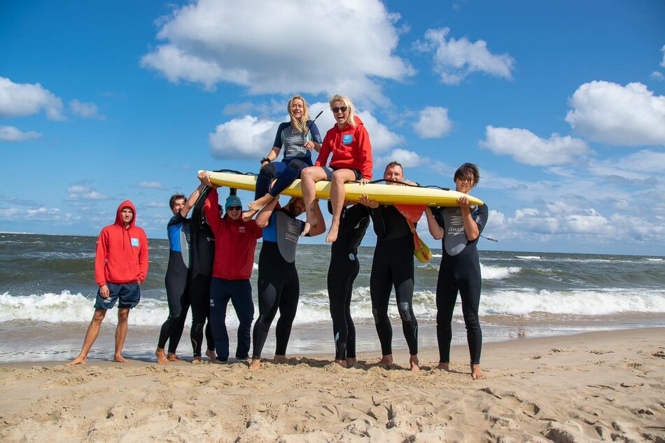 kilkunastu ratowników i ratowniczek na plaży, dwie młode kobiety siedzą na desce windsurfingowej, pozostałe osoby trzymają deskę z nimi nad głowami