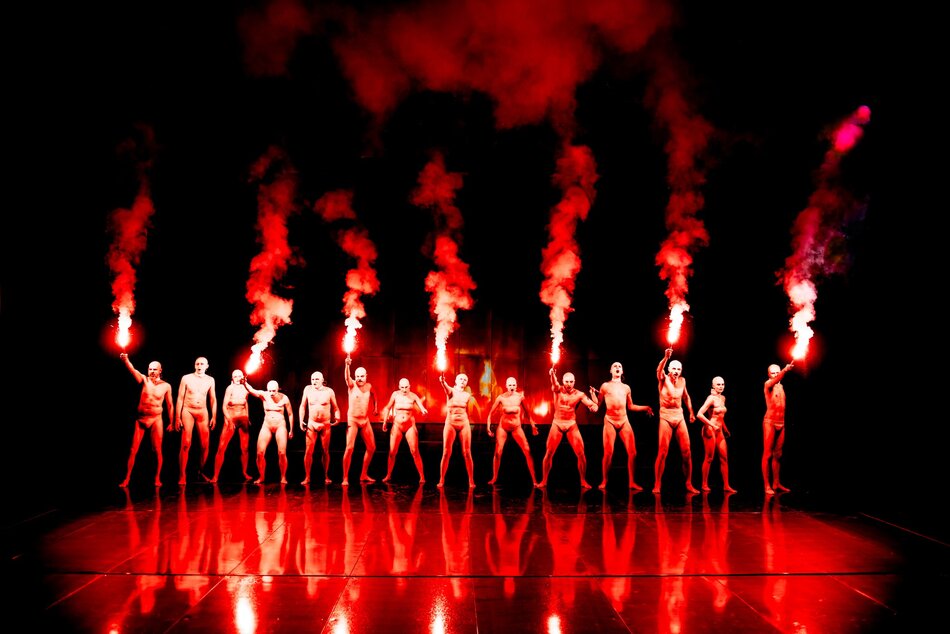 Kilkunastu aktorów z czerwonymi racami stoi na scenie, ucharakteryzowani są na nagich