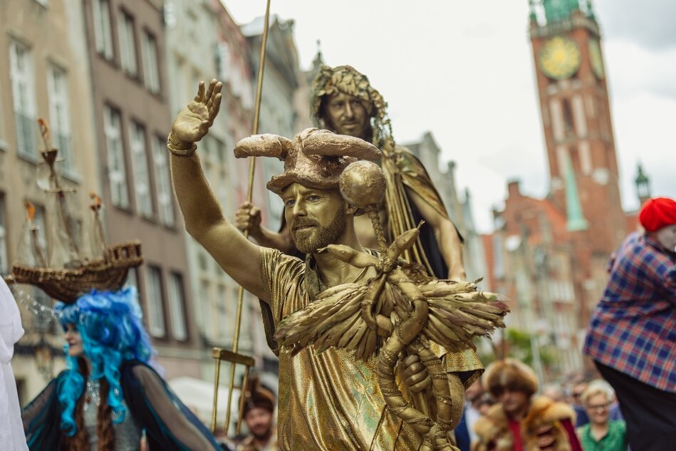 Aktor pomalowany na złoto i w złotym stroju grający postać Neptuna, trzyma w ręku trójząb. Idzie w pochodzi