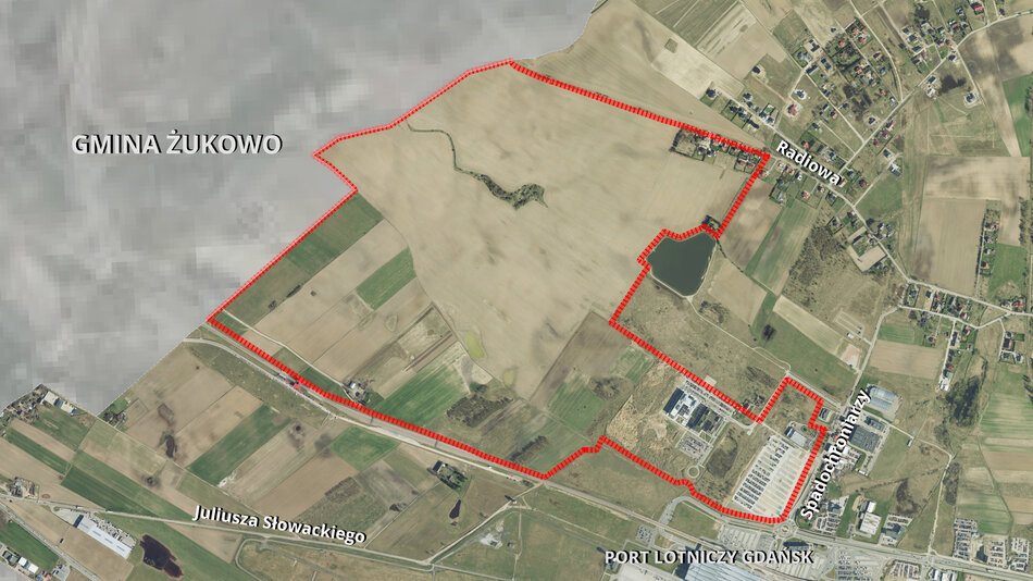 Zdjęcie przedstawia widok z lotu ptaka na określony obszar zaznaczony czerwoną linia przerywaną. Na dole i w górnej części obrazu znajdują się nazwy miejsc, takie jak Juliusza Słowackiego i Radiowa. Wzdłuż dolnej krawędzi zdjęcia widoczna jest nazwa Port Lotniczy Gdańsk, na lewej stronie widoczny jest napis GMINA ŻUKOWO, co sugeruje, że ten obszar należy do określonej jednostki administracyjnej w Polsce. 