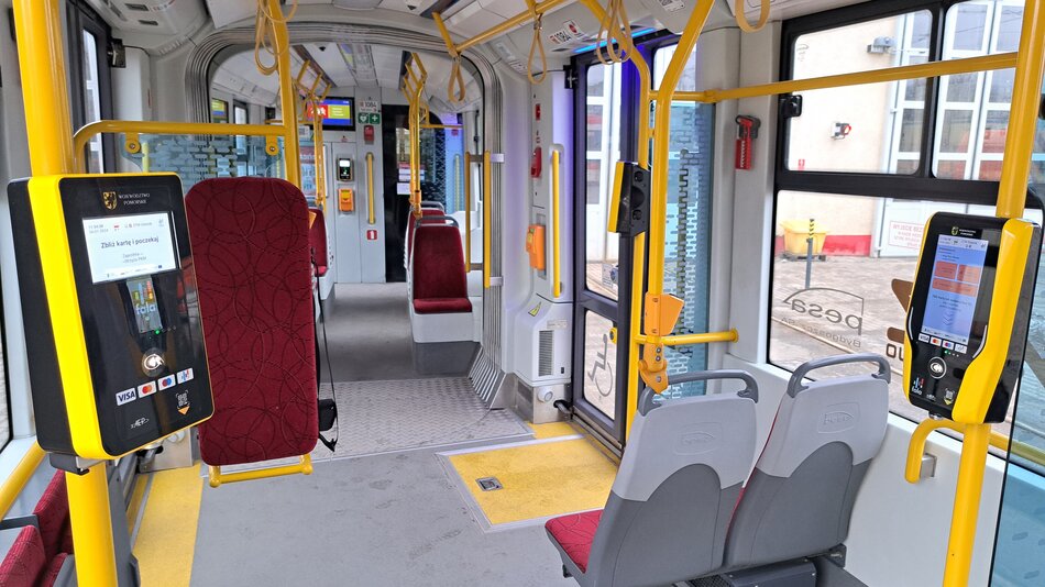 Na zdjęciu widzimy wnętrze współczesnego tramwaju lub autobusu. Po lewej stronie znajduje się urządzenie do płatności bezgotówkowych oraz terminal biletowy z ekranem dotykowym. Wyświetlacz terminala pokazuje informacje po polsku, co może wskazywać na to, że pojazd znajduje się w Polsce. Pojazd jest wyposażony w czerwone siedzenia z wzorzystym obiciem oraz szare plastikowe oparcia. Na podłodze widać żółtą wykładzinę w miejscu przeznaczonym dla osób niepełnosprawnych lub wózków. Po prawej stronie jest drugi taki sam terminal biletowy. Wnętrze jest czyste i wydaje się być nowoczesne, z metalowymi poręczami i żółtymi uchwytami dla pasażerów stojących. Na zewnątrz widać budynek, prawdopodobnie przystanek lub zajezdnię