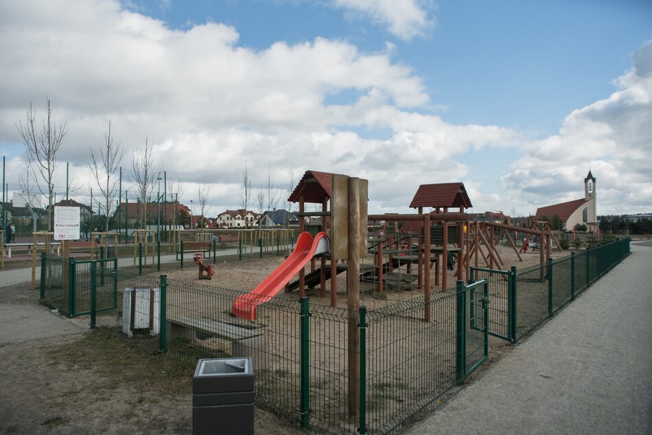 na zdjęciu ogrodzony metalowym ogrodzeniem plac zabaw, widać tam drewniane elementy zabawowe