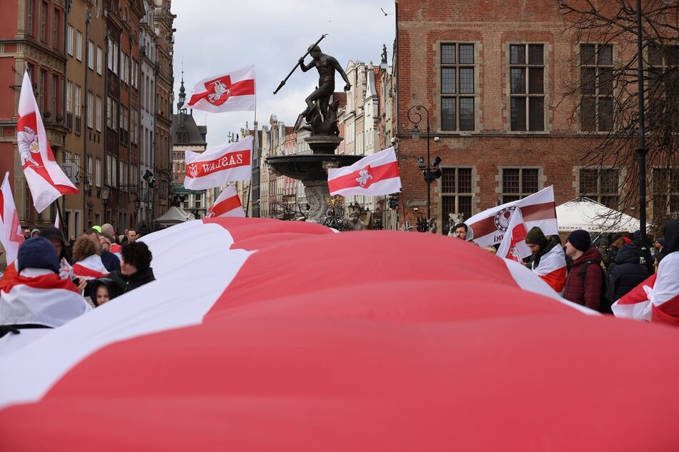Na zdjęciu widać grupę ludzi trzymających duży czerwono-białą flagę na tle miejskiego krajobrazu. W tle stoi Fontanna Neptuna, może to być postać historyczna lub bohater wojenny. Wokół pomnika i nad tłumem unoszą się również flagi Białorusi. 