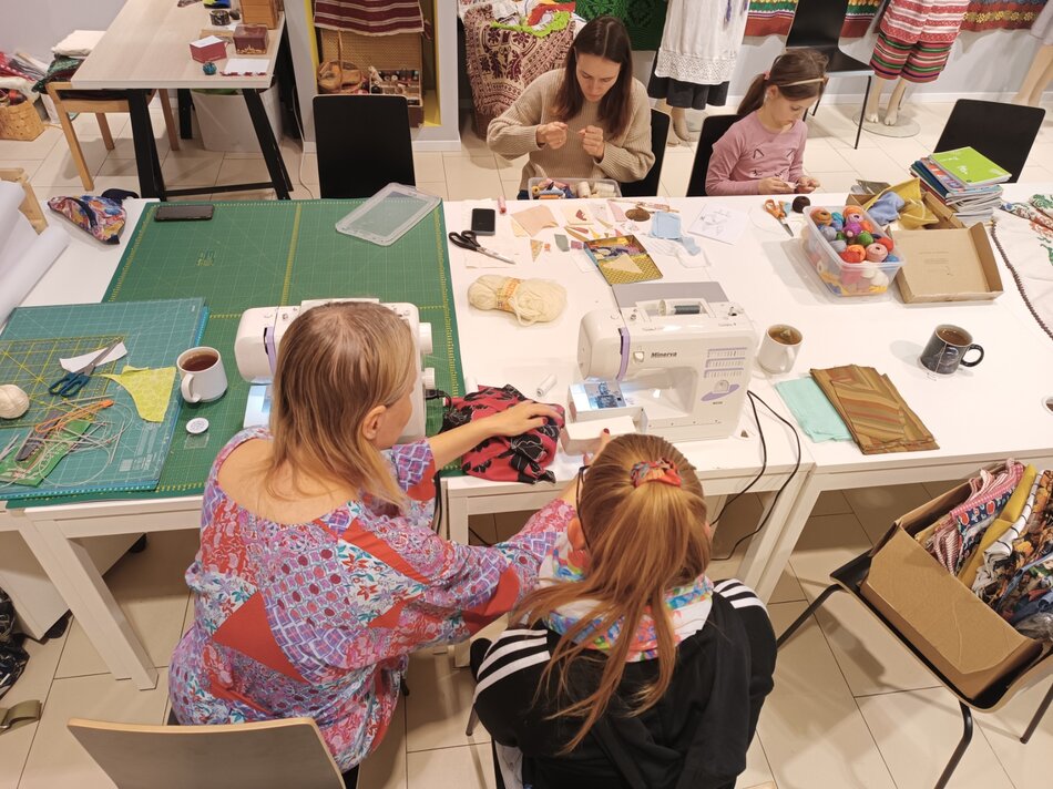 na zdjęciu duży biały stół, na nim stoi kilka maszyn do szycia fragmenty materiałów, po obu stronach stołu siedzą dorosłe kobiety i małe dziewczynki