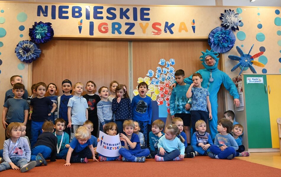 grupa dzieci w wieku ok 7 lat pozuje w sali szkolnej pod napisem Niebieskie Igrzyska, jest z nimi kilkoro dorosłych, w tym mężczyzna w niebieskim przebraniu, wiele dzieci w grupie także jest ubranych na niebiesko