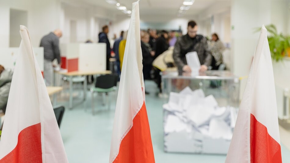 To zdjęcie przedstawia wnętrze pomieszczenia, w którym prawdopodobnie odbywają się wybory. Na pierwszym planie widzimy dwie flagi z białym i czerwonym pasem – barwy te mogą sugerować, że to wydarzenie ma miejsce w Polsce. W tle znajdują się osoby oddające głosy do przezroczystych urn wyborczych. Wszyscy uczestnicy wydają się być skupieni na procesie głosowania. Pomieszczenie jest jasne i nowoczesne, z meblami w prostym stylu