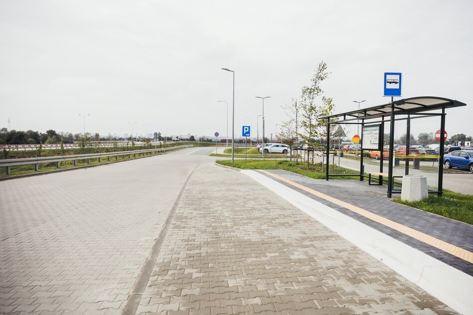 na zdjęciu fragment jezdni o nawierzchni z kostki betonowej, po prawej wiata przystankowa, w tle kilkanaście zaparkowanych samochodów