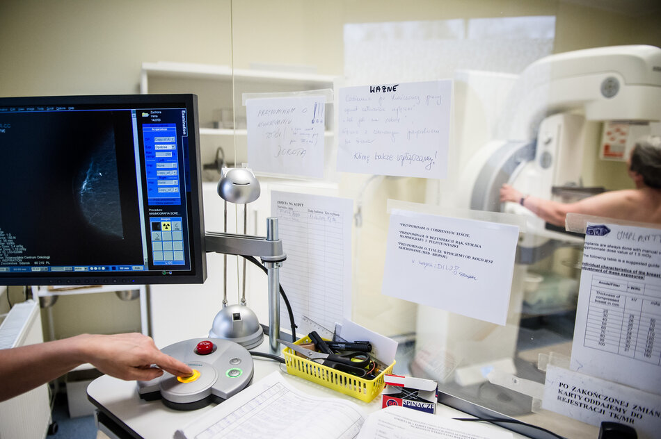 Z lewej strony ekran monitora w gabinecie medycznym z prawej w tle urządzenie do badania piersi i fragment stojącej nagiej kobiety w średnim wieku