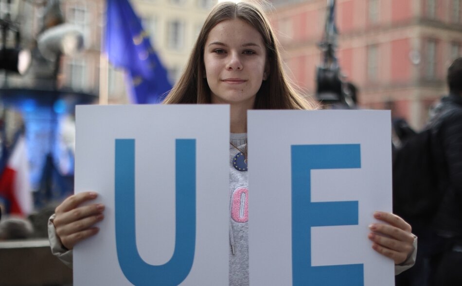 Na zdjęciu widzimy młodą kobietę, która trzyma dwa białe plakaty z literami U i E w kolorze niebieskim, które razem tworzą skrót UE, co jest powszechnie używanym skrótem dla Unii Europejskiej. Kobieta ma długie, proste brązowe włosy i jest ubrana w szary top. Stoi na tle zatłoczonego miejsca, prawdopodobnie na jakimś zgromadzeniu lub demonstracji, co sugerują nieostre tło i widoczne flagi. Na jej twarzy maluje się delikatny uśmiech.